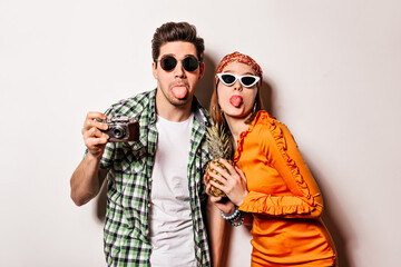 Cheerful man and woman in sunglasses are showing tongues and posing with retro camera and pineapple on isolated background