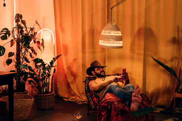 relaxed man with cowboy hat lying down on a lounge at home holding telephone and typing on the...