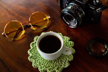 Xícara com café, câmera antiga e óculos com lentes amarelas sobre uma mesa de madeira.