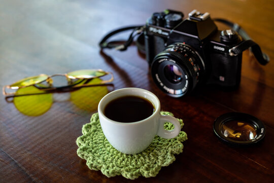 Xícara com café, câmera antiga e óculos com lentes amarelas sobre uma mesa de madeira.