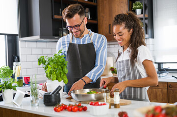 Young cheerful multi-ethnic couple preparing tomato pasta together at home kitchen