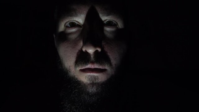 A mysterious aggressive man in the darkness. Violence and horror concept. Low light close-up studio shot on dramatic male face. Selective focus on the face. Frightening bearded criminal in the night.
