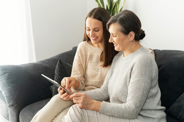 Side view an adult daughter and a senior mother spend time with a tablet indoor, a young woman showing to mature mom smth in a network, helps to figure out with a device - Powered by Adobe