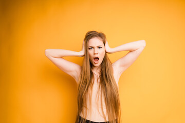 Attractive Caucasian girl with long hair holds her head with her hands and shouts, on an orange background. Emotional woman