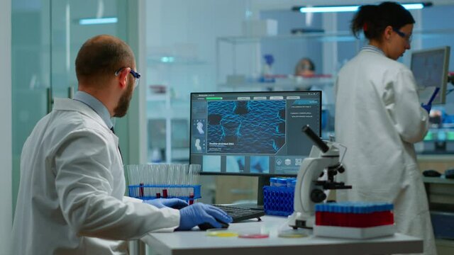 Man biologist checking DNA informations typing on pc in modern equipped laboratory. Scientists examining vaccine evolution in medical lab using high tech, chemistry tools for scientific research.