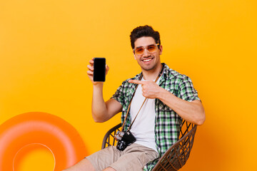 Charming man in summer outfit points to black smartphone. Cheerful guy posing on chair with retro camera on orange background with inflatable circle