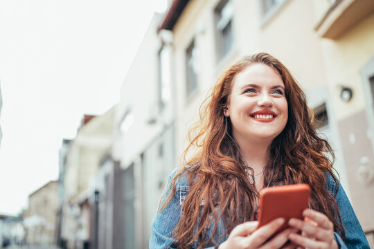 Smiling red curly long hair caucasian teen girl walking on the street and browsing the internet using the modern smartphone. Modern people with technology devices concept image.