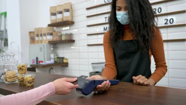 Multiracial black barista girl in uniform and medical mask and protective gloves gives a visitor coffee in a disposable cardboard glass and dessert in a paper bag to go. Coffee to go concept