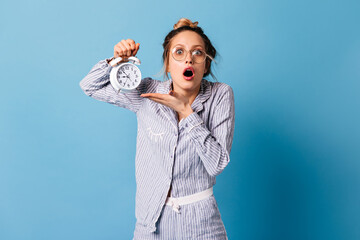 Awakened girl in glasses and blue sleepwear points at ringing alarm clock and stares into camera in...