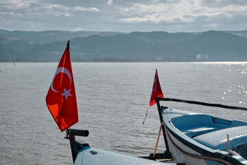 Fototapeta premium Small and blue boats near the coast of Lake of Uluabat in Turkey. Small Turkish flag on the boat with lake and mountain background. Bursa. Golyazi (Apollyon). Bursa, Turkey. 22.01.2021.