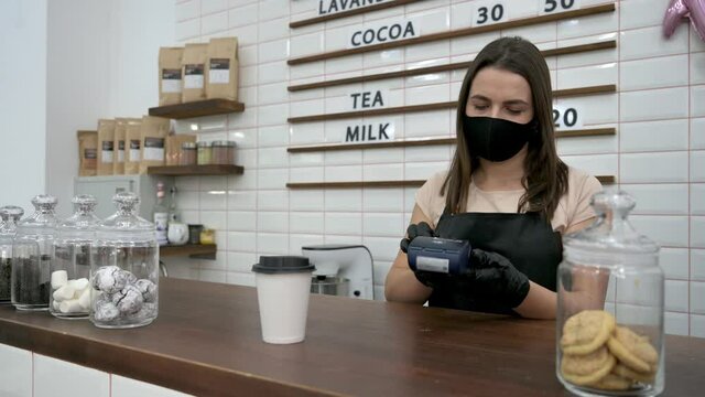 A friendly caucasian waitress, wearing medical mask giving disposable cardboard cup of coffee and terminal for cashless payment to a visitor, standing inside coffee shop
