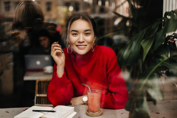 Portrait of joyful lady in red oversized sweater holding glass of freshly squeezed watermelon juice, sitting in cafe against background of plants