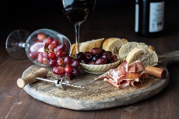 Charcuterie board with olives, prosciutto and bread and a tipped over wine glass filled with red...