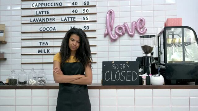 Frustrated emotional African American waitress, barista or small business owner in a black apron, stands inside coffee shop, restaurant or bar and shows at a signboard SORRY CLOSED