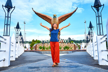 Happy girl tourist posing next to sculpture of a red eagle spreading its wings. Popular tourist spot on Langkawi island - obrazy, fototapety, plakaty