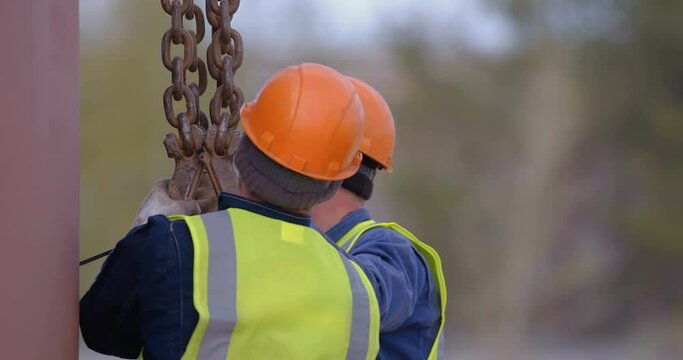 Builders in orange helmets are about to lift a load with a crane and takes hooks on metal chains by hands
