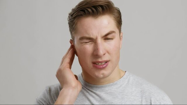 Otitis. Portrait of young man suffering from ear pain, close up, grey background
