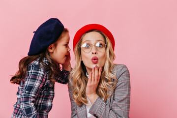 Mom and daughter gossip on pink background. Little girl in beret tells secret to astonished woman