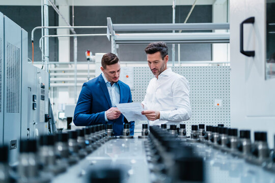 Male professionals discussing over document while standing by manufacturing equipment in factory