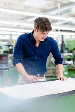Young businessman with machine part looking at floor plan on desk in industry