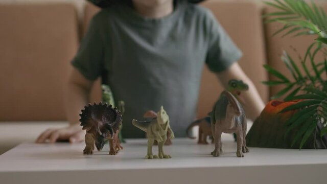 A boy plays with dinosaur figurines on the table. The little explorer in the hat. Handheld close up shot.