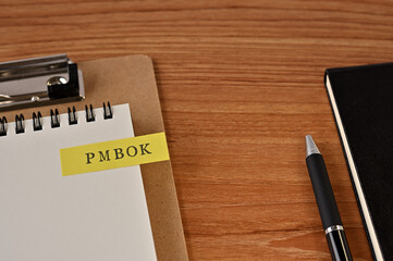 There is a notebook, a pen, and a clipboard with a sticky note stuck to it that says PMBOK written on it. It was an abbreviation for Project Management Body of Knowledge.