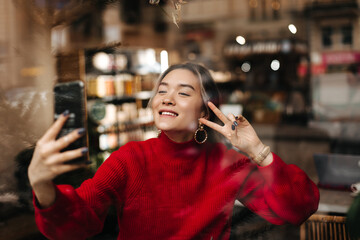 Gorgeous woman in massive earrings and stylish bright sweater takes selfie and shows peace sign, enjoying rest in cafe