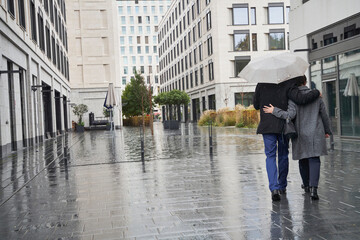 Businesswoman and senior businessman with arms around walking under umbrella in city during rainy season