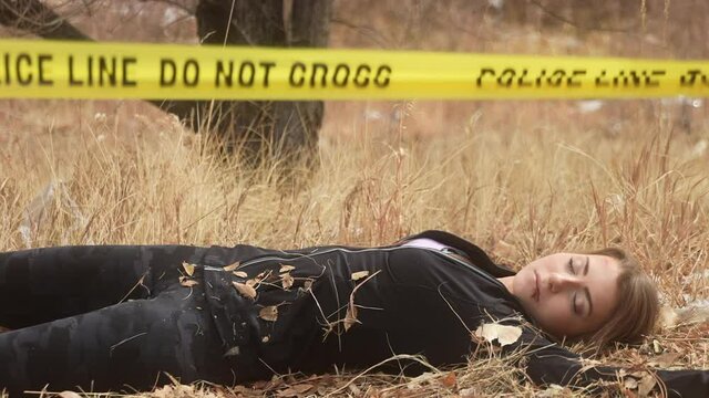 Detective approaches body of deceased female jogger and examines corpse for evidence behind police tape on grass field