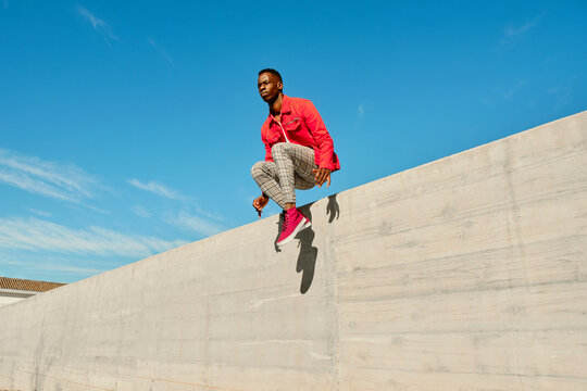 Young man in red jacket and checked pants jumping from concrete wall