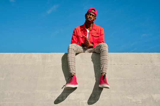 Portrait of young man in red jacket and baseball cap sitting on concrete wall