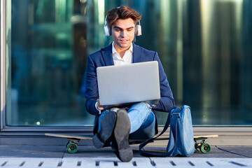 Young businessman working on laptop while listening music sitting on skateboard during sunny day