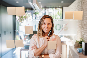Smiling businesswoman looking at adhesive notes stuck on glass wall in office