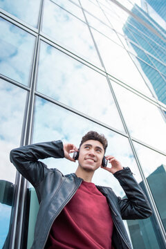 Happy young man listening music through headphones against glass building