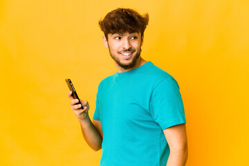 Young indian man holding a phone looks aside smiling, cheerful and pleasant.
