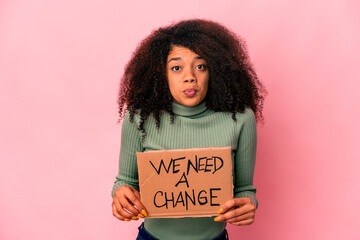Young african american curly woman holding a we need a change cardboard shrugs shoulders and open...