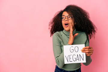 Young african american curly woman holding a go vegan placard is saying a secret hot braking news and looking aside