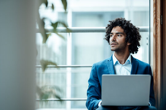 Thoughtful male professional looking away while sitting with laptop against window at workplace