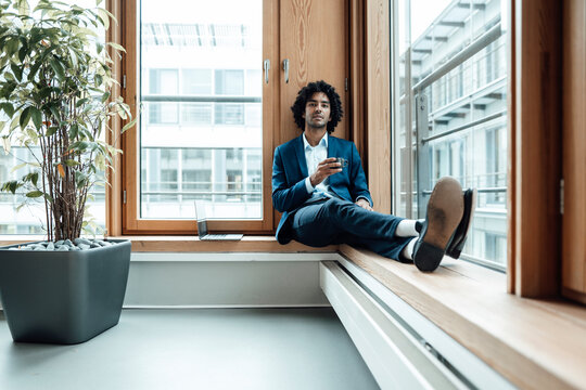 Confident male entrepreneur holding coffee cup while sitting against window at office