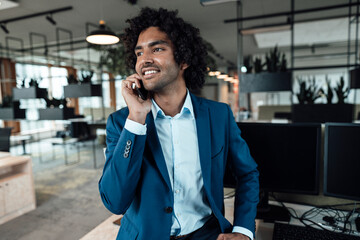 Smiling young businessman talking on smart phone while looking away at workplace