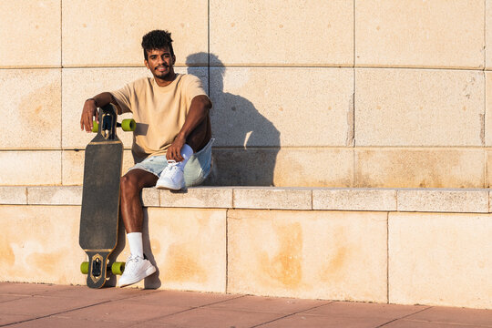 Smiling young man with skateboard sitting on retaining wall during sunset