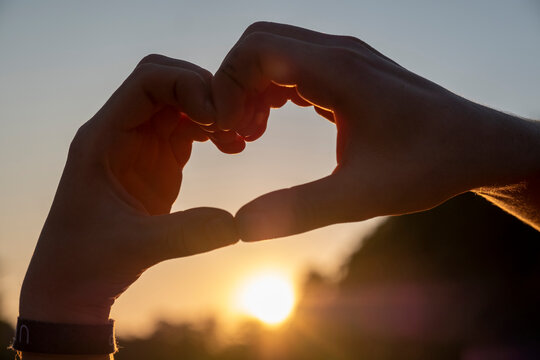 Man making heart shape with hands against sky during sunset