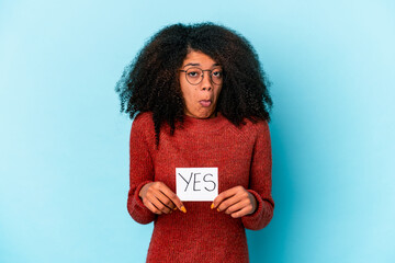 Young african american curly woman holding a yes placard shrugs shoulders and open eyes confused.