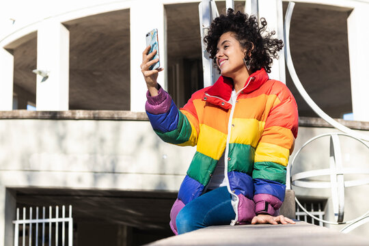 Woman wearing colorful jacket smiling while taking selfie through mobile phone sitting against building