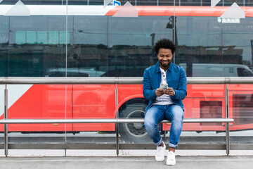 Smiling stylish mid adult man using mobile phone while sitting at bus stop