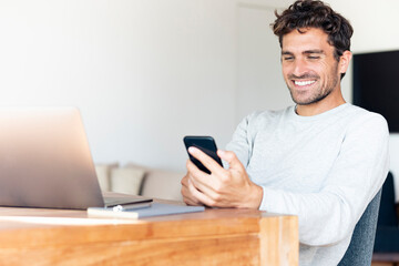 Smiling man with laptop using mobile phone while sitting by table at home