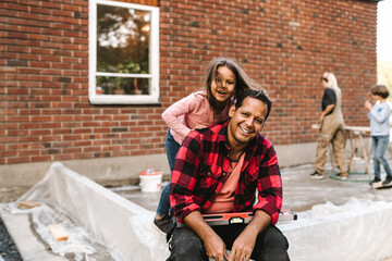 Portrait of smiling confident man with daughter sitting on concrete wall against house