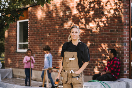 Portrait of confident woman with drill machine while family in background against brick wall
