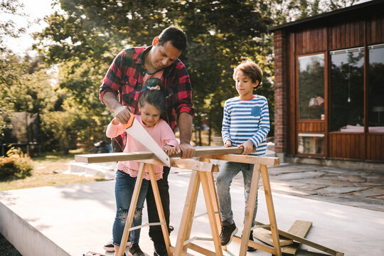 Daughter and father using hand saw on wood while son standing by table outside house