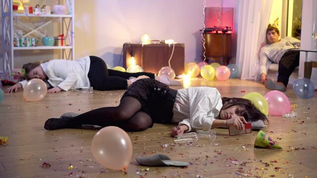 Wide shot of slim young woman sleeping lying on party floor checking smartphone. Portrait of drunk Caucasian millennials resting after partying indoors. Youth lifestyle and alcohol abuse concept.